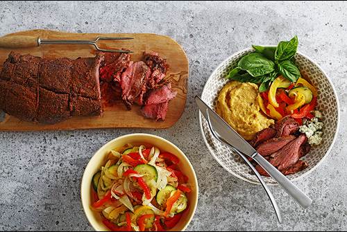 Balsamic beef tenderloin on a cutting board with vegetables and polenta served in dishes