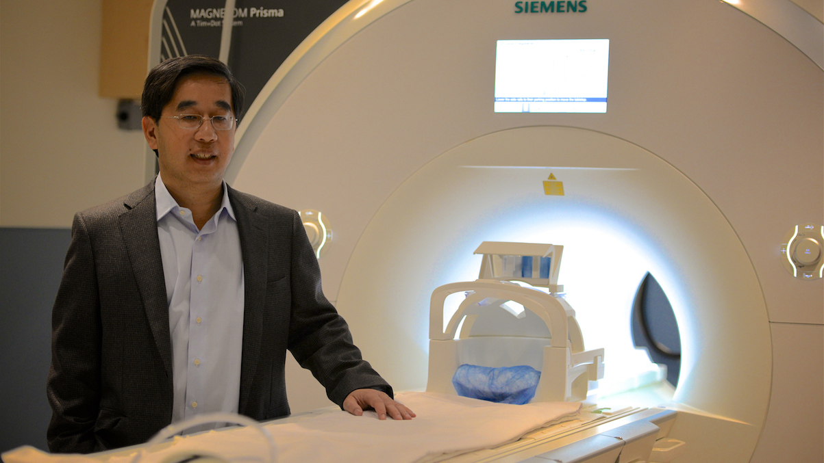 Dr. Jack Tu stands next to a CT Scanner wearing a grey blazer and a blue button down shirt. 