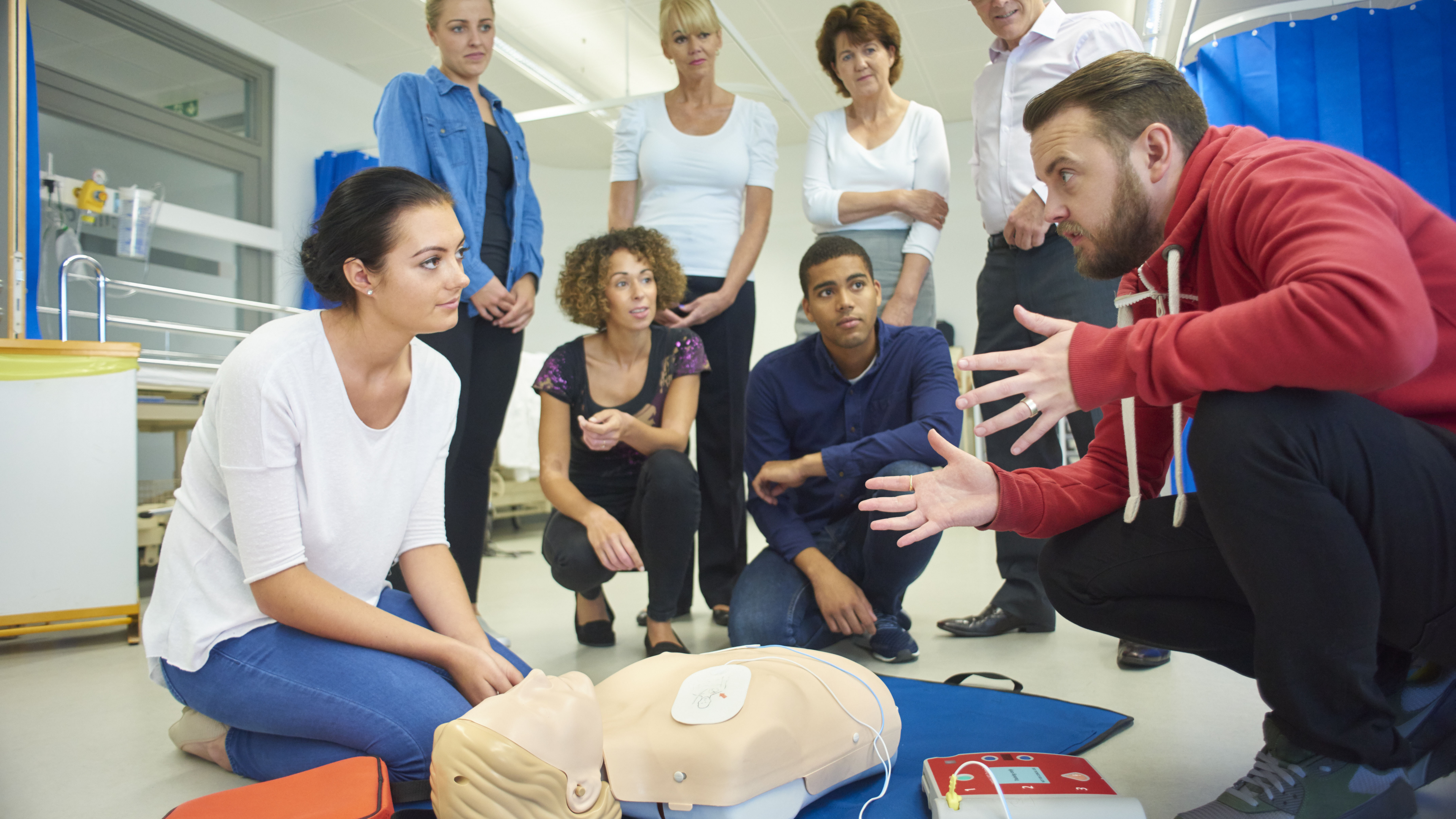 a mixed age group listen to their tutor as he shows the procedure involved to resuscitate using a defibrillator