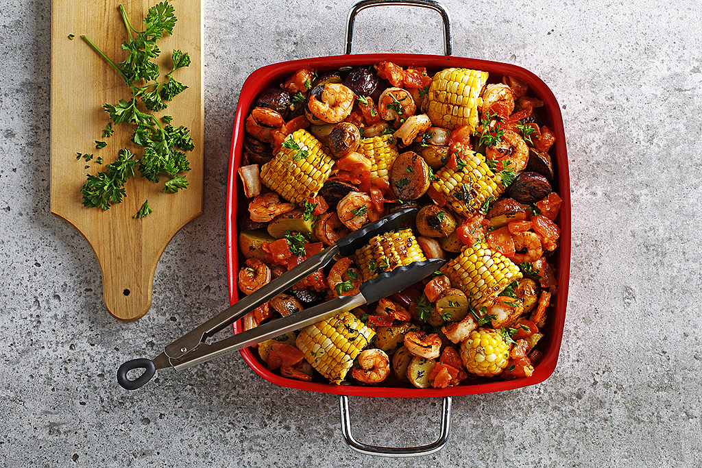 Grilled shrimp and corn casserole in a square red serving dish with tongs