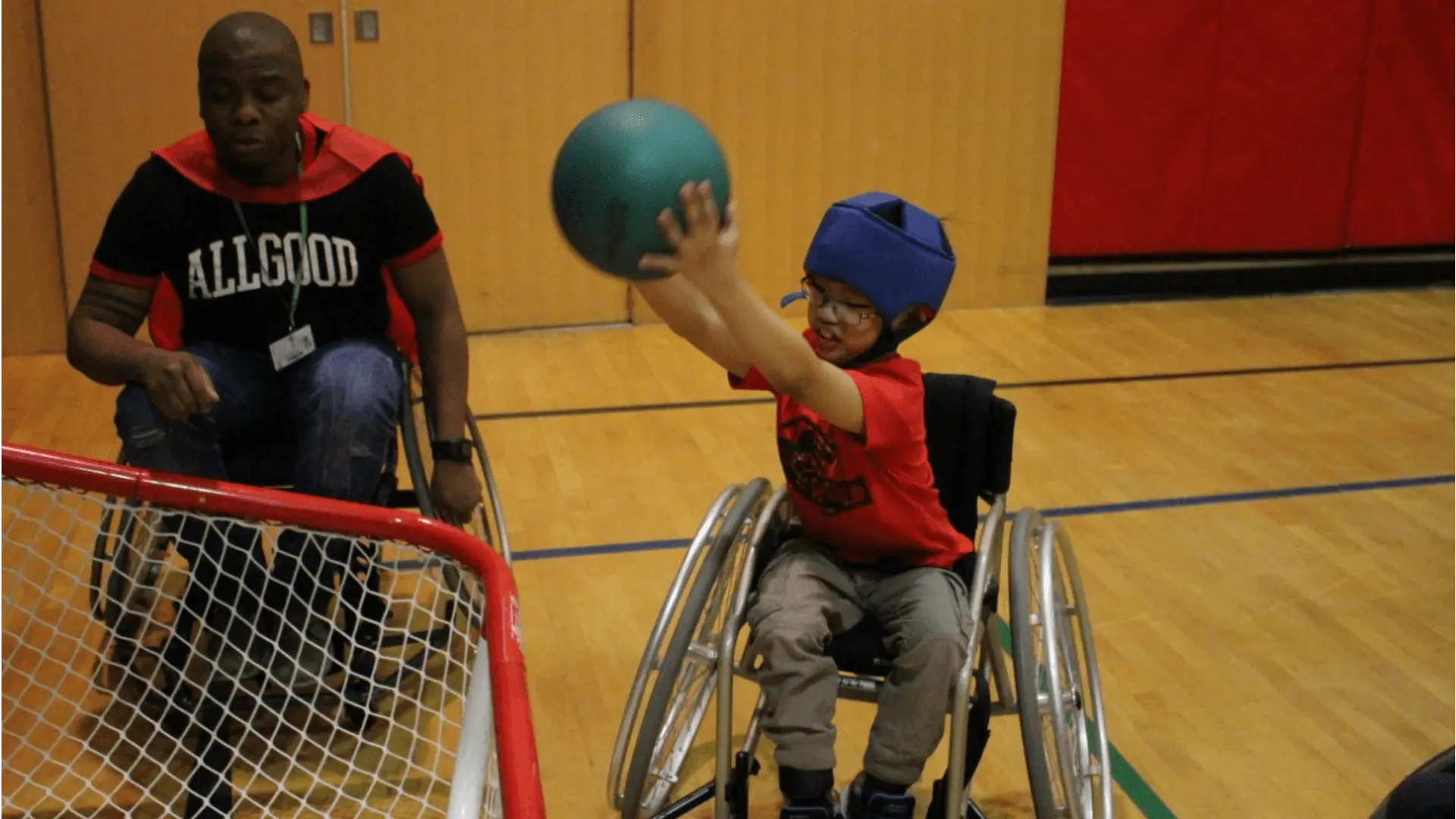 A student in a wheelchair lifts a ball over his head aiming for a net in a school gymnasium.