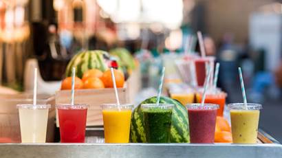 Six clear plastic cups full of colourful juices sit in front of various fruits.