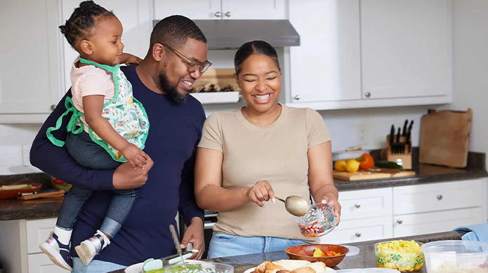 A family of three by the kitchen table, with the mother preparing the meal and the father holding their child in one arm.