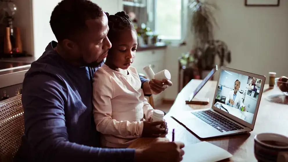 A father with his daughter on his lap attending a virtual doctor’s appointment. 