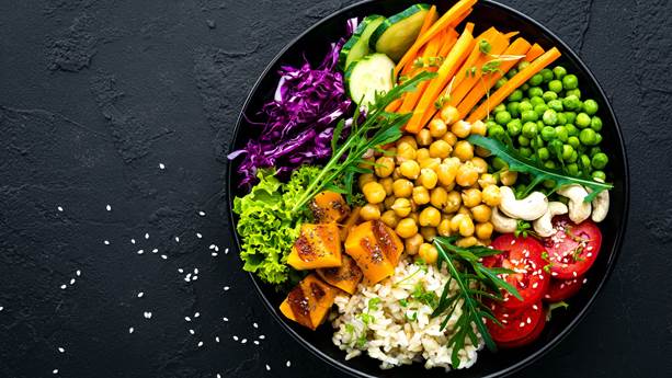 A large bowl on a dark background overflows with vegetables including chickpeas, carrots, cabbage and tomatoes.