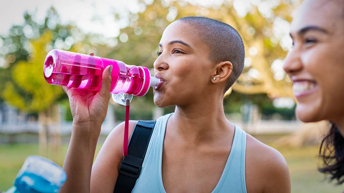 Young woman sipping water from a pink water bottle after working out at a park with friends. 
