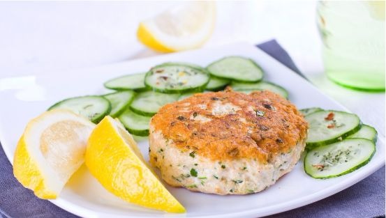 Salmon patty on plate with sliced cucumber and lemon wedges