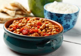 Cooked chickpeas, tomatoes in ceramic pot. Bowl of white rice and pita in background