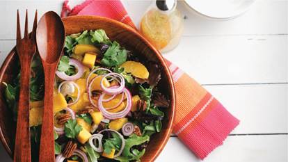 Mixed greens, mango and pecan salad in a wooden bowl with wooden utensils.