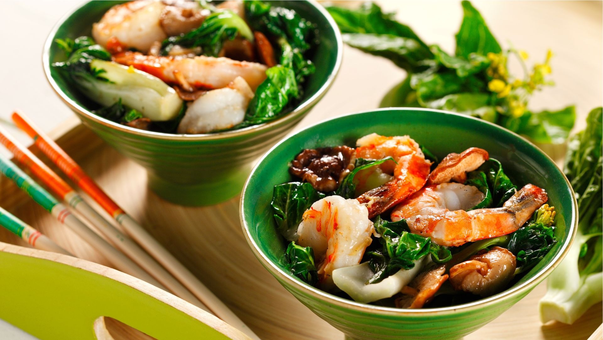 Two bowls filled with shrimp, mushrooms and bok choy on wooden serving tray with chop sticks