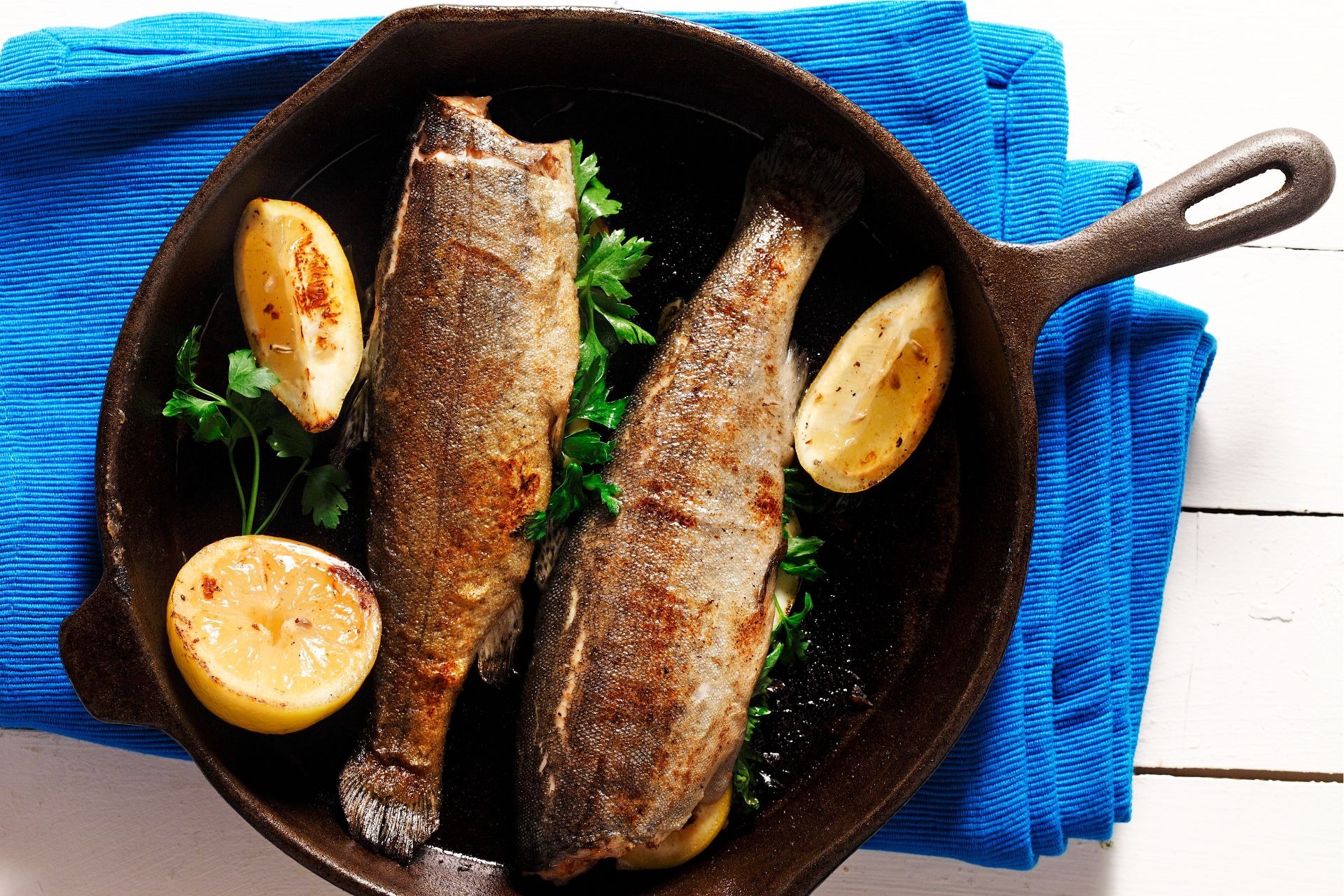 Two pieces of trout in cast iron skillet with sliced lemon and fresh parsley.