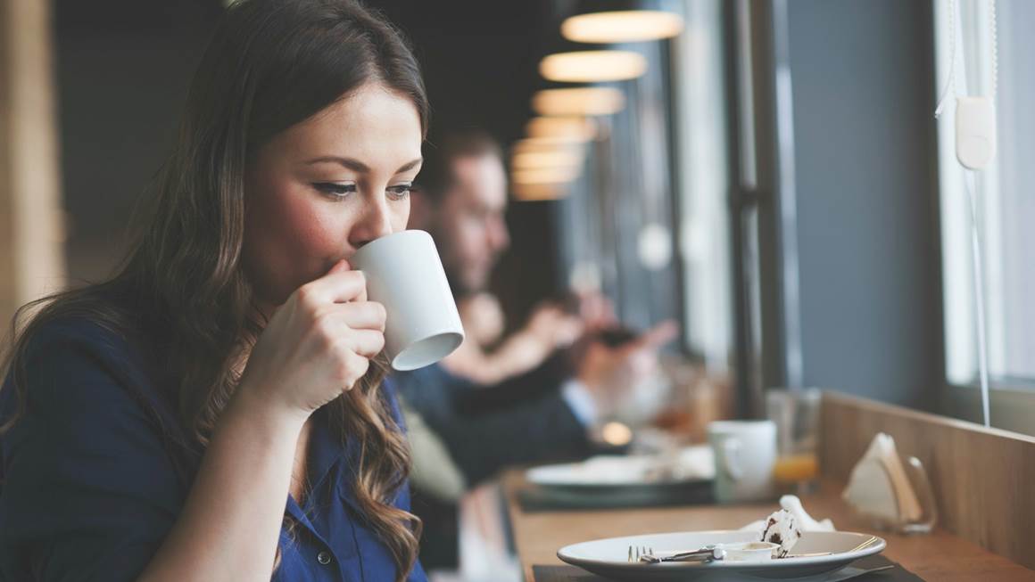 Woman sitting in a coffee shop sips from a white mug.