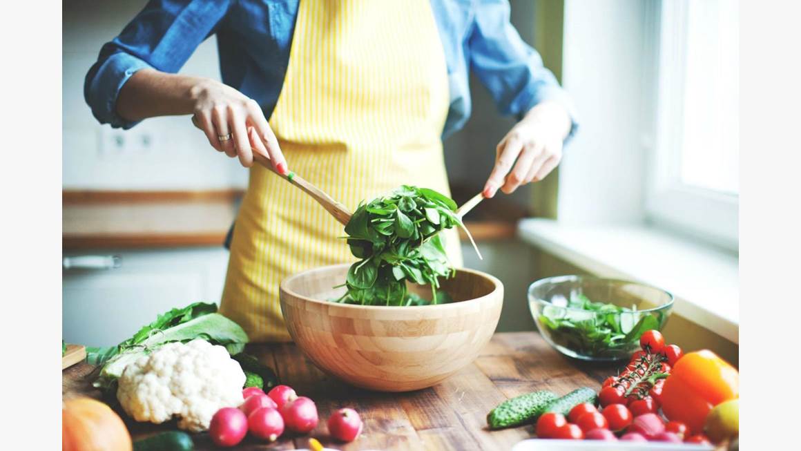A woman wearing a yellow apron tosses green salad in a wooden bowl.