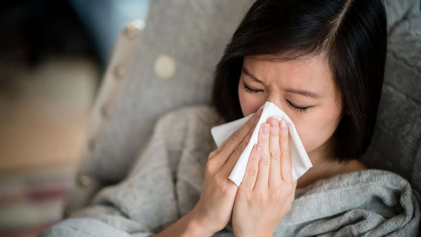 A woman wearing a gray sweater blows her nose into a tissue.
