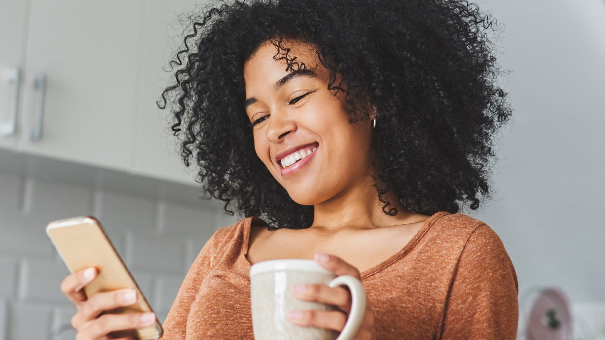A woman smiling down at her phone with a cream mug in her hand