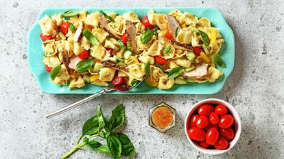 Chicken tortellini pasta salad on a blue serving platter with bowl of cherry tomatoes, dressing and a basil sprig on the side.