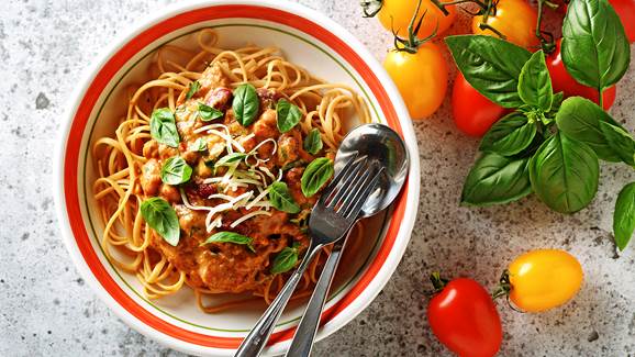 Roasted tomato sauce linguini in a serving bowl with silverware and whole tomatoes and basil clippings on the side. 