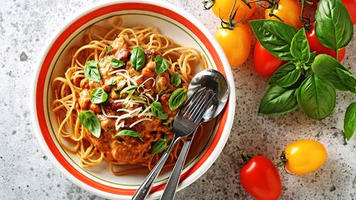 Roasted tomato sauce linguini in a serving bowl with silverware and whole tomatoes and basil clippings on the side. 