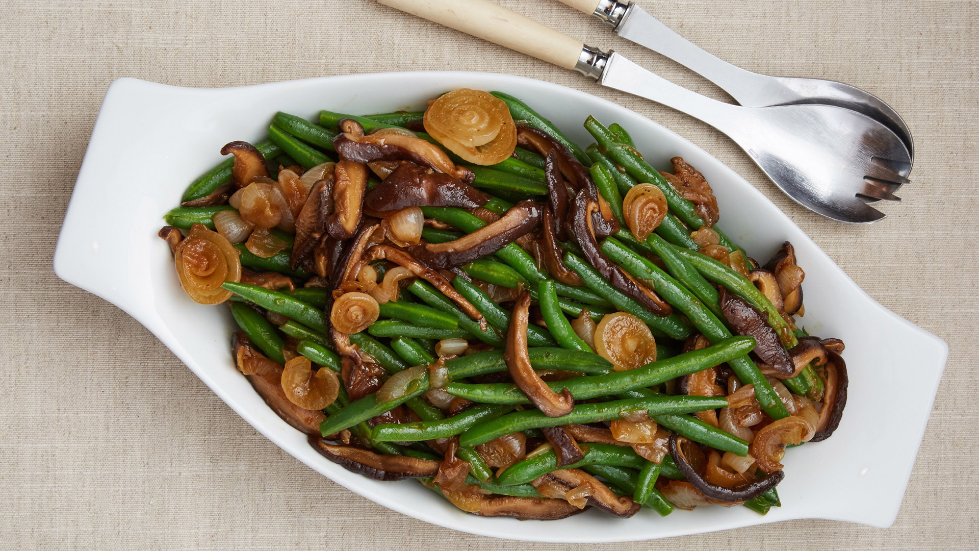 Asian green beans with wild mushrooms and Cipollini onions in a white serving dish and fork and spoon. 