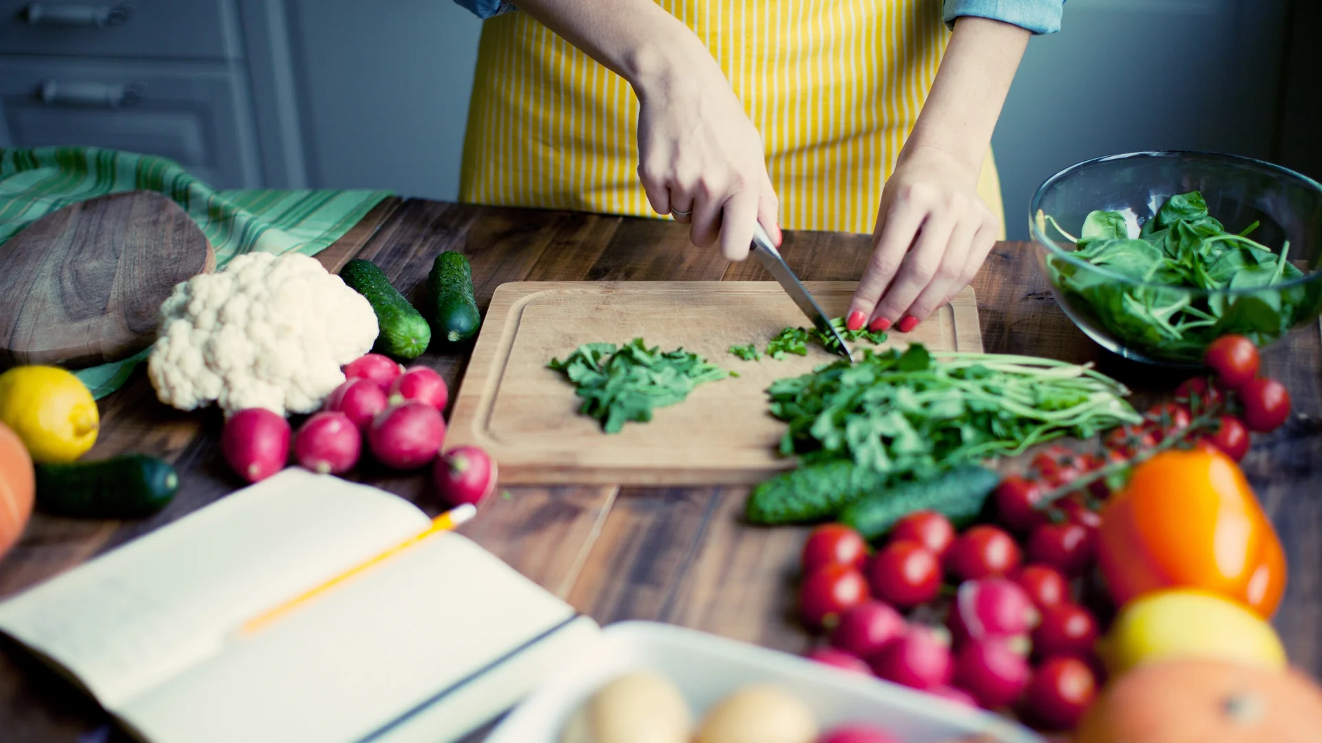 A person is chopping vegetables on a cutting board, with a wide variety of fresh vegetables and an open recipe book on the table.