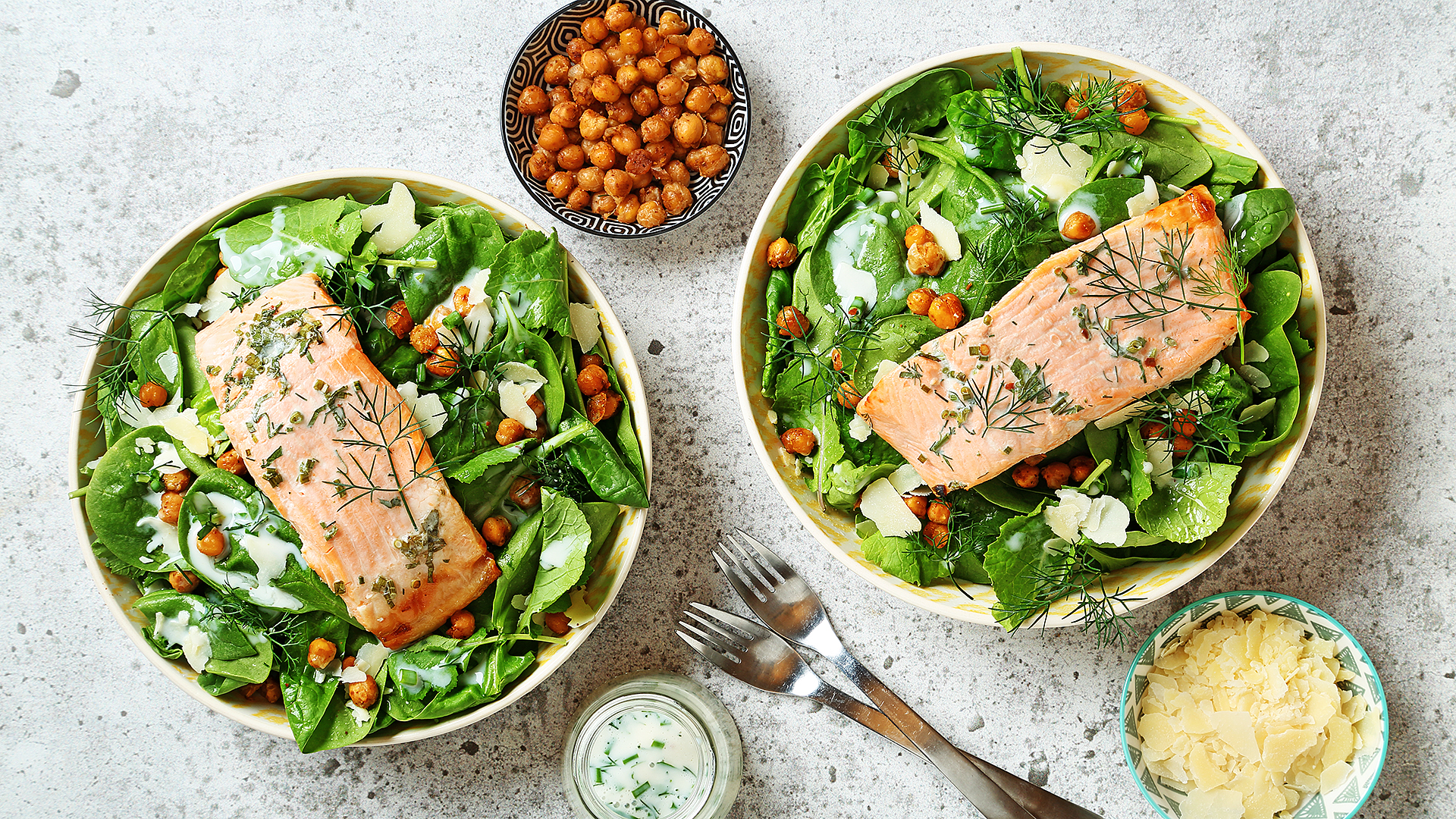 Kale chickpea salad with trout with silver utensils and bowls of chickpeas, dressing and parmesan cheese on the side.