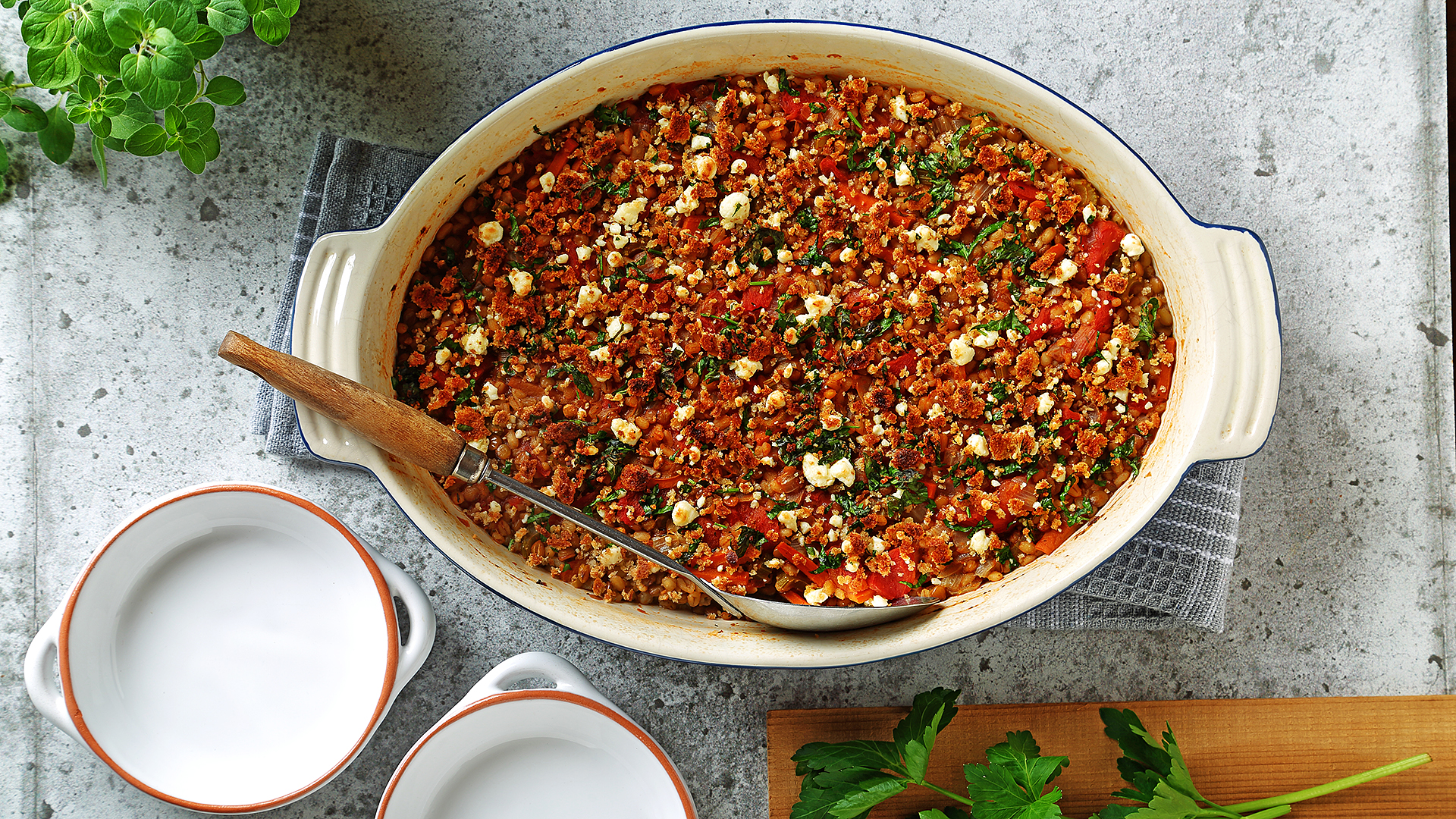 Lentil and barley oven bake casserole in ceramic serving dish with a serving spoon and two empty bowls.