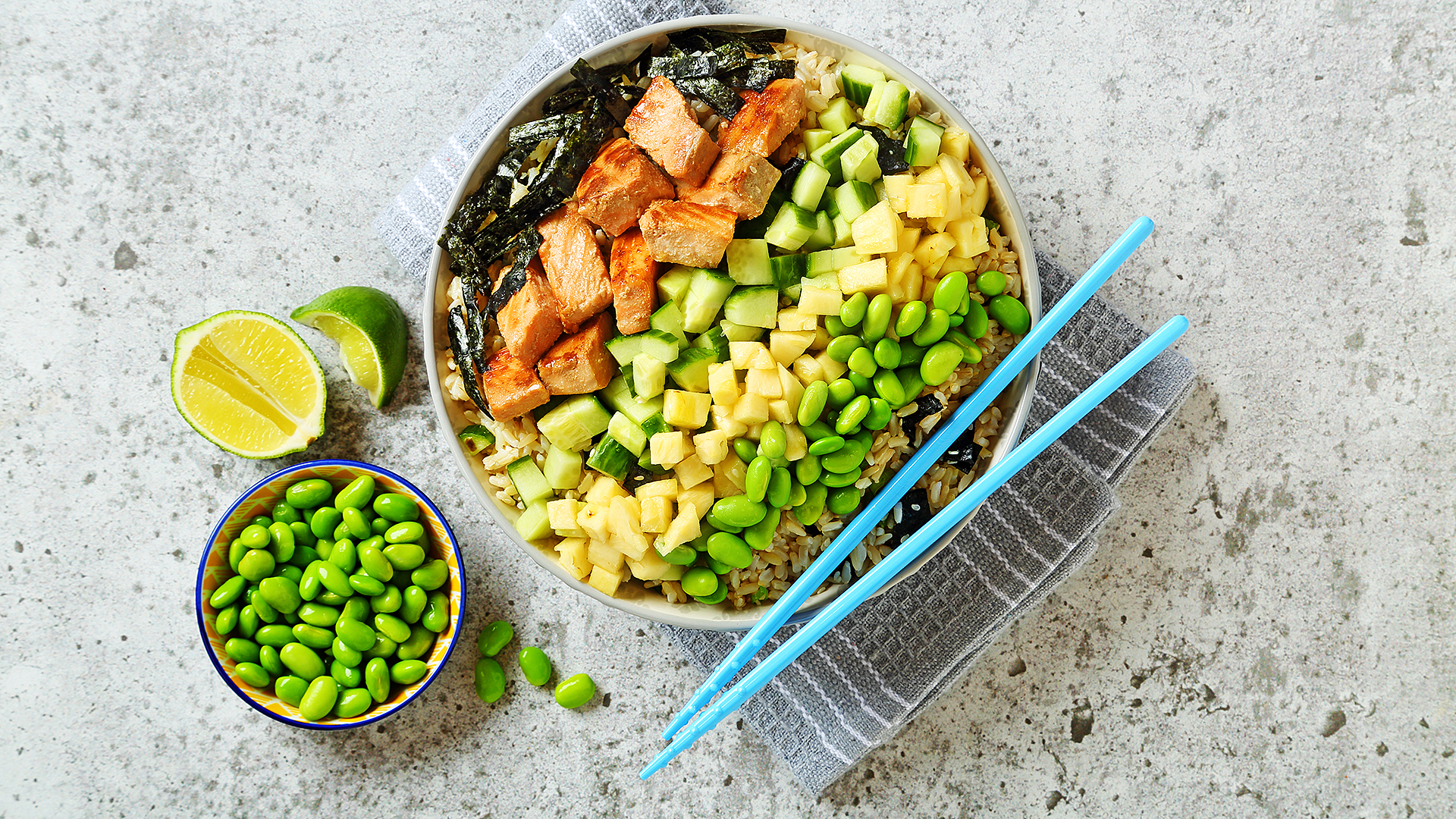 Salmon poke style salad in a bowl with blue chopsticks and edamame and lime on the side