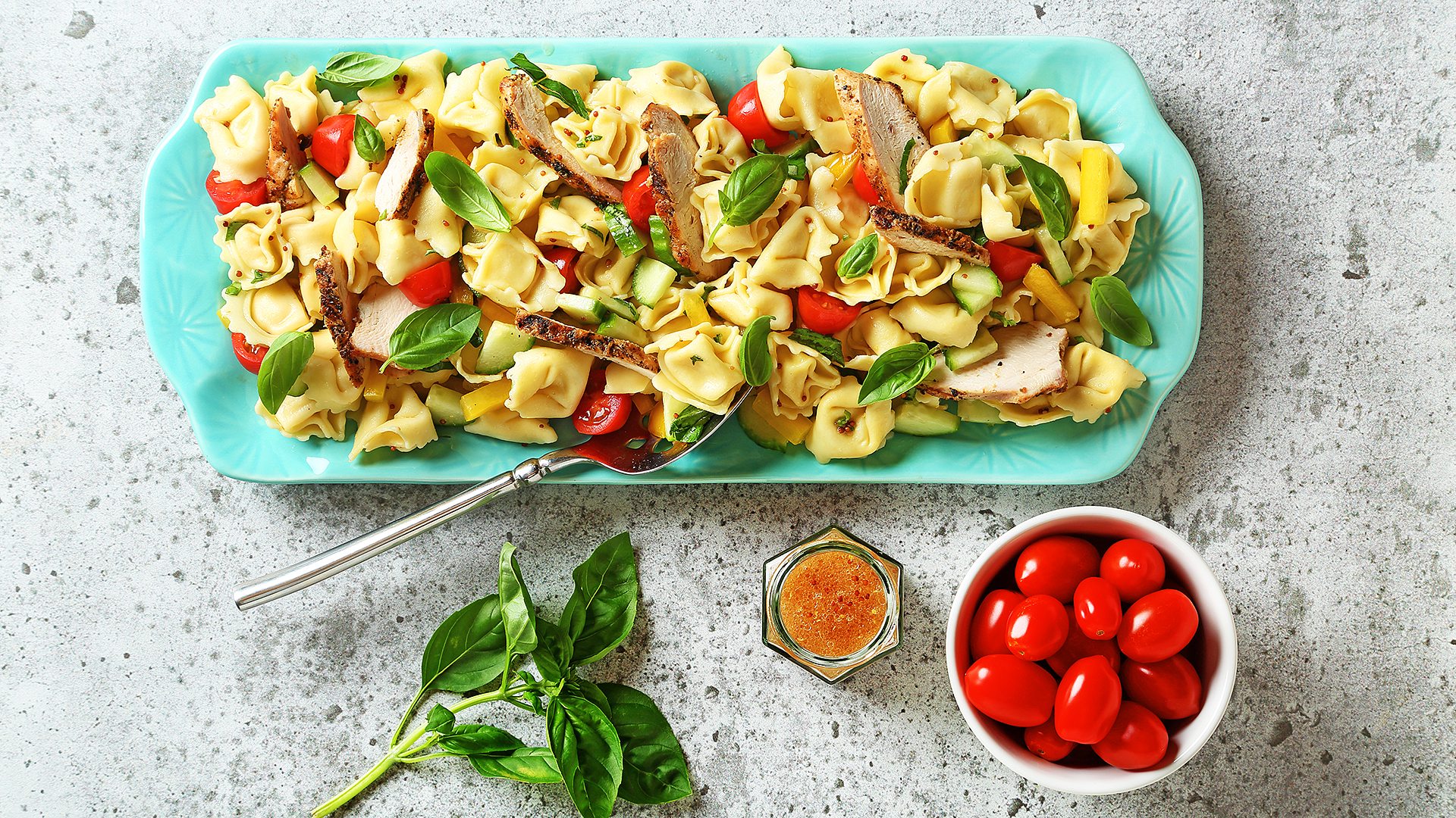 Mediterranean tortellini pasta salad on a blue serving platter with bowl of cherry tomatoes, dressing and a basil sprig on the side.