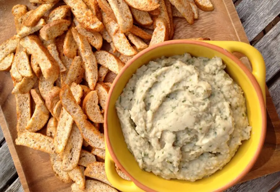 A bowl of navy bean hummus next to a pile of seasoned bagel crisps on a wooden tray.