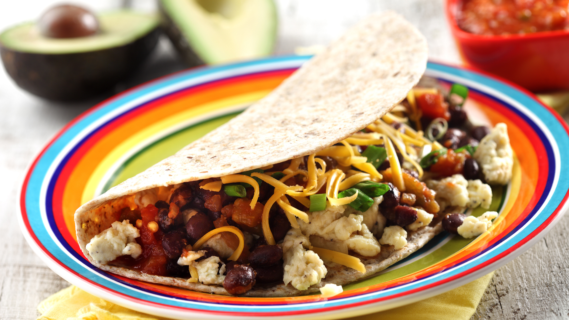 Breakfast taco on a rainbow coloured plate on top of a yellow napkin with a halved avocado in the background.