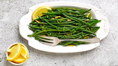 Blistered green beans with ginger on a white serving platter with silver serving fork and lemon slices on the side. 