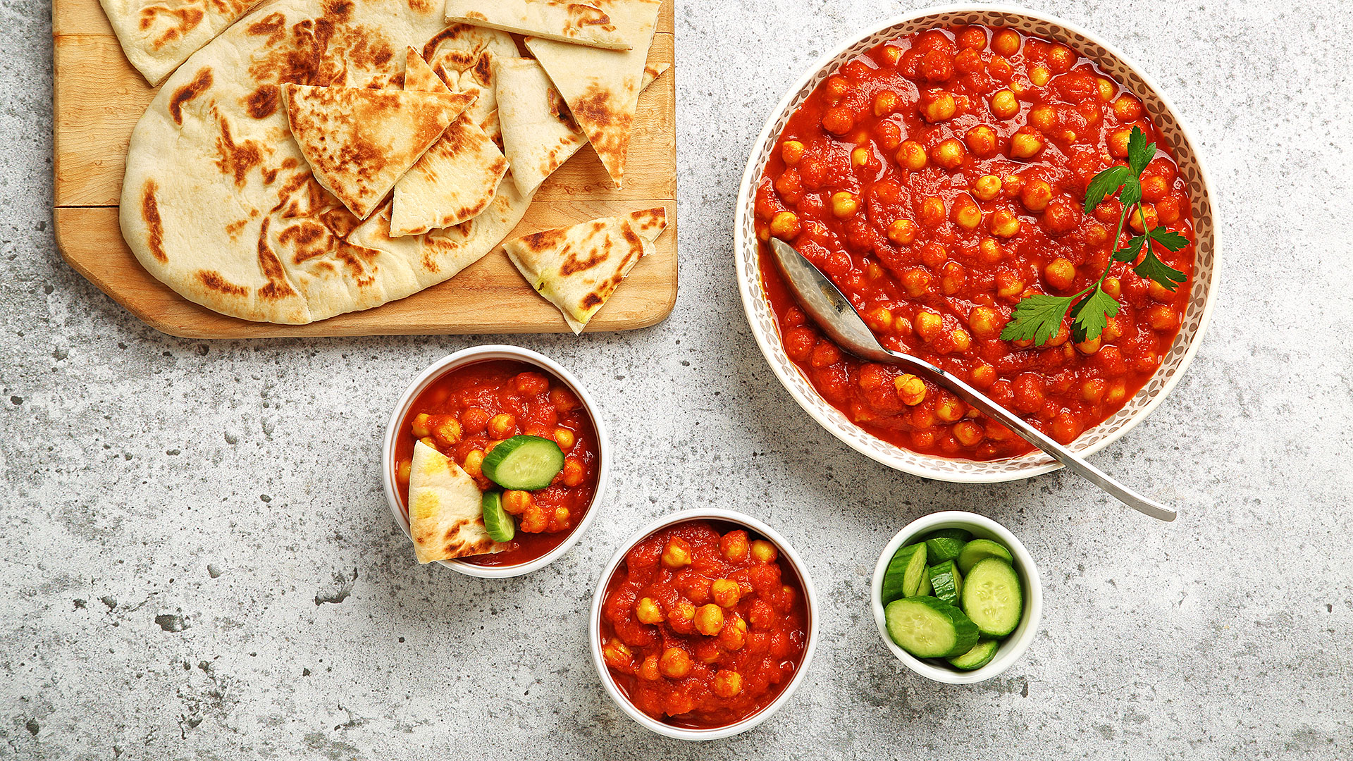 Garlic tomato chickpeas in a big serving bowl with naan on a wooden cutting board and cucumber slices in a small bowl on the side.