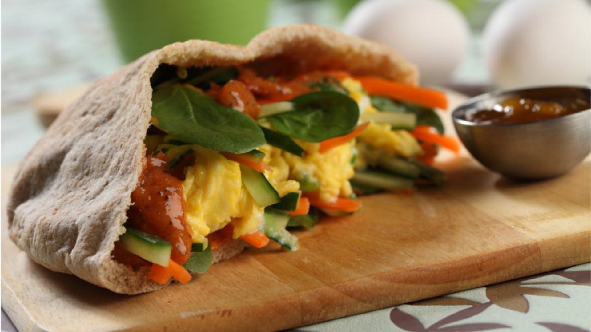 A whole wheat pita cut in half and filled with vegetables and curried egg, displayed on a cutting board and a side of sauce.