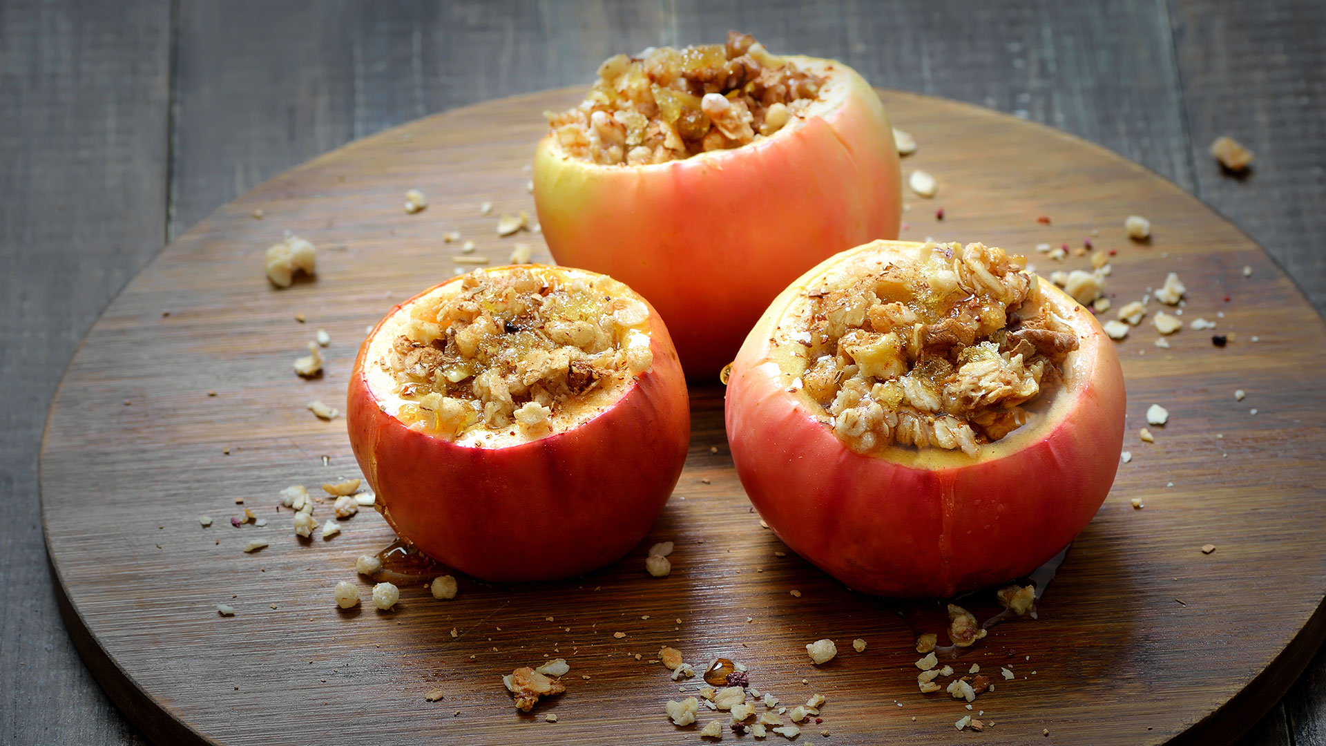 Three baked red apples on a wooden board with granola filling.