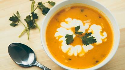 Carrot squash soup with cream and a parsley sprig in a white bowl on a wooden board next to a metal spoon.