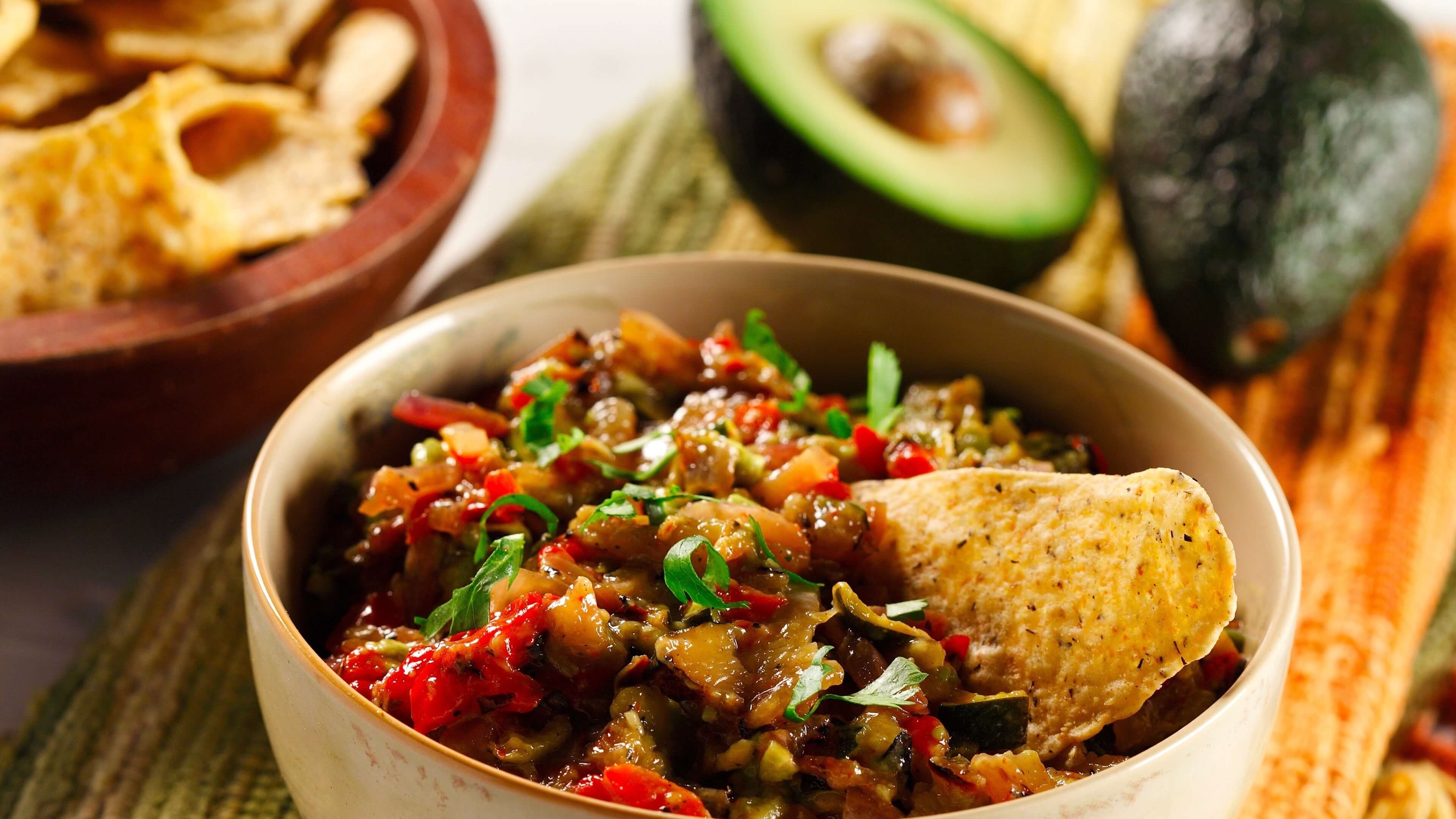 Vegetable guacamole in a bowl with tortilla chips and avocado in background