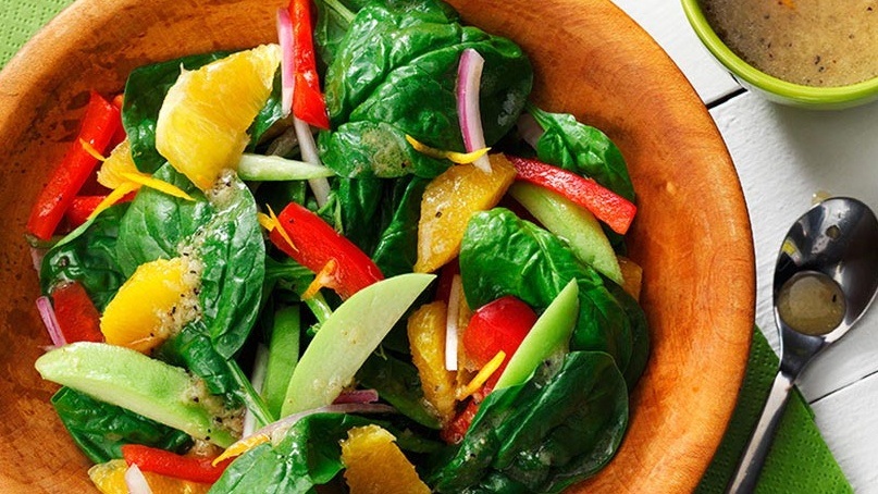 Bowl of spinach with orange segments, red peppers, onion in wooden bowl with vinaigrette in background