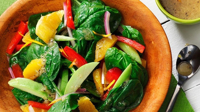 Bowl of spinach with orange segments, red peppers, onion in wooden bowl with vinaigrette in background