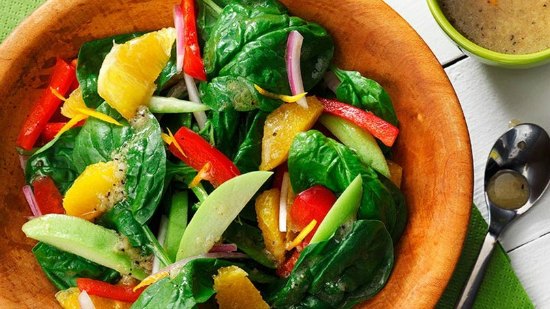 Bowl of spinach with orange segments, red peppers, onion in wooden bowl with vinaigrette in background 