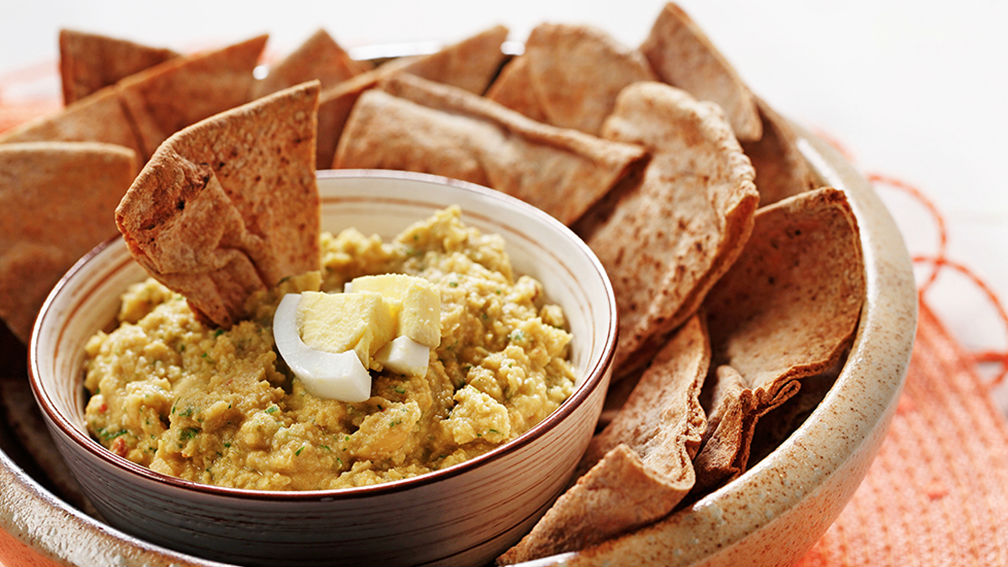 Bowl of hummus with sliced hard boiled egg on top sitting inside larger bowl filled with whole wheat pita bread.
