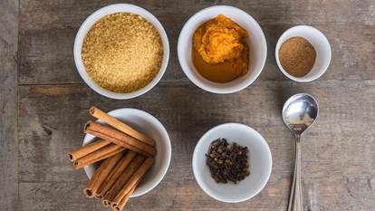 Pumpkin spice ingredients in white bowls and a serving spoon on a wooden tabletop. 