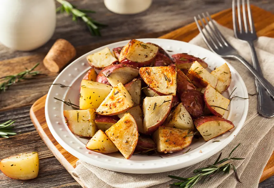 Roasted potatoes with rosemary on a white plate, placed on a wooden board with cutlery.