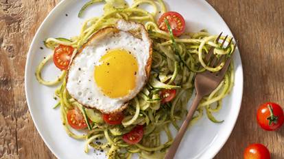 Zucchini noodles with cherry tomatoes,  pesto sauce and a sunny side up egg on a white plate and bronze fork. 
