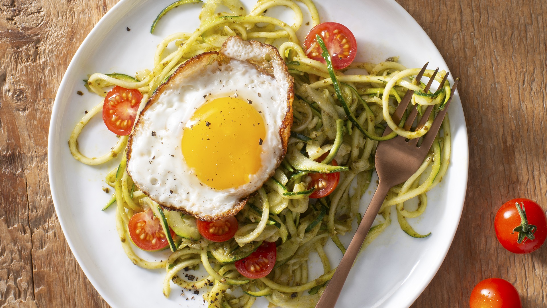 Zucchini noodles with cherry tomatoes,  pesto sauce and a sunny side up egg on a white plate and bronze fork.
