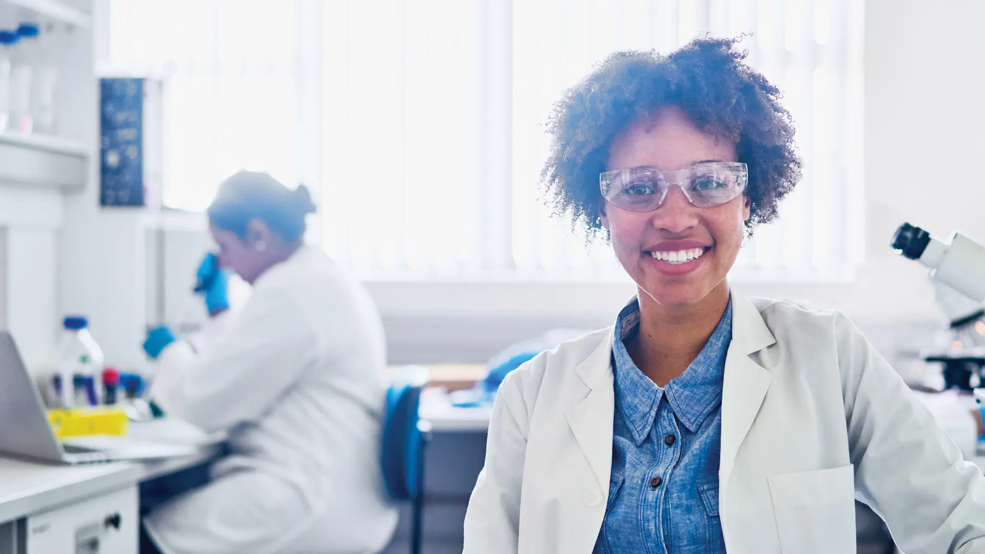 A woman researcher smiles while in a lab, with another researcher working in the background.