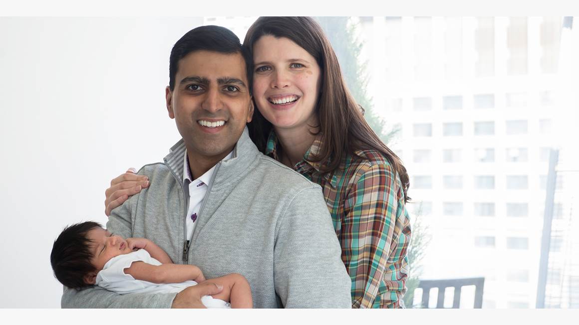 Irfhan Rawji and his wife Christine, stand close together with arms wrapped around their new born baby boy, Zain, while smiling at the camera. 