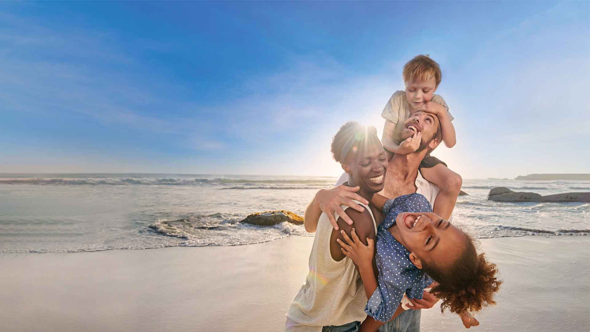 A family of 4 of mixed ethnicity laughing happily on the beach.