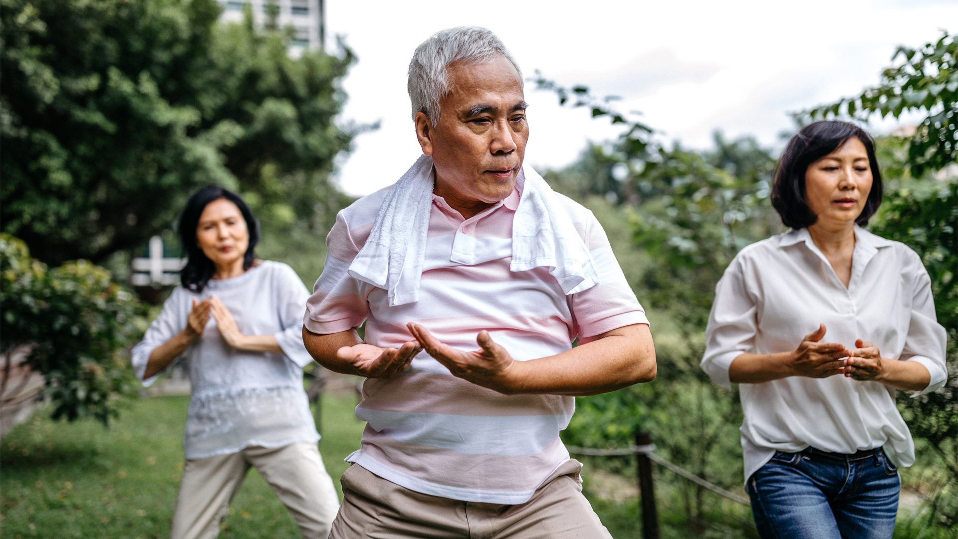A group of people practicing tai chi in a park, standing with their hands raised. 