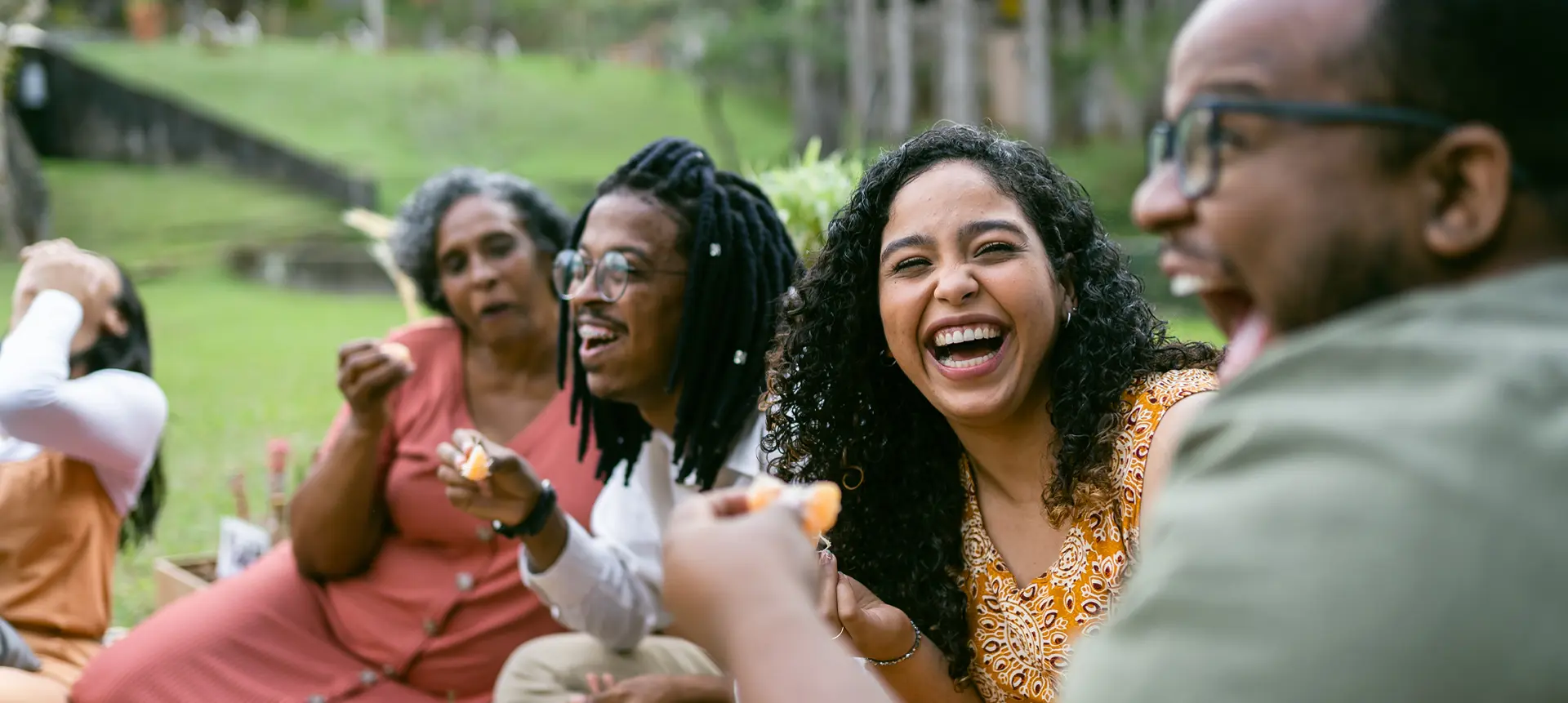 Group of people sitting outdoors on grass, sharing food and socializing at a picnic. 