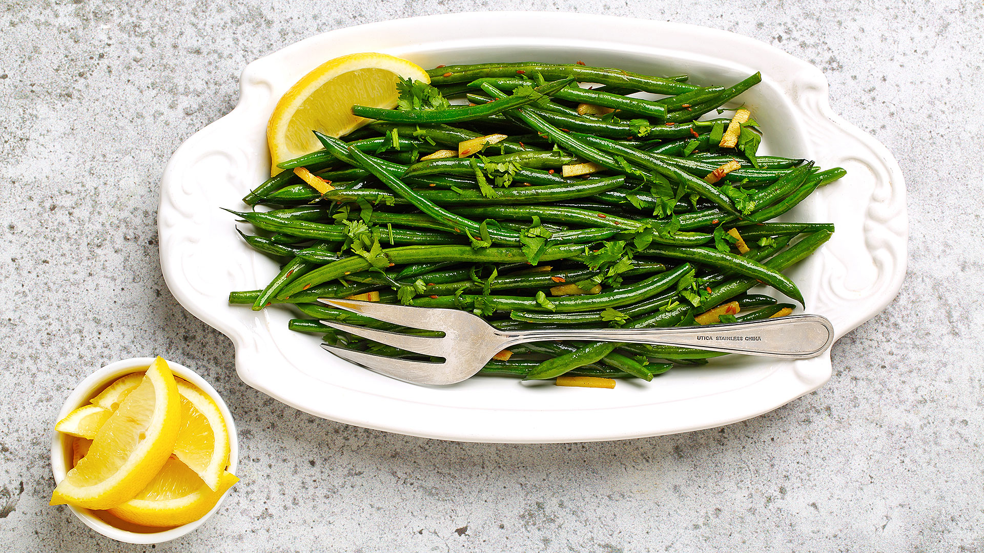 Blistered green beans with ginger on a white serving platter with silver serving fork and lemon slices on the side. 